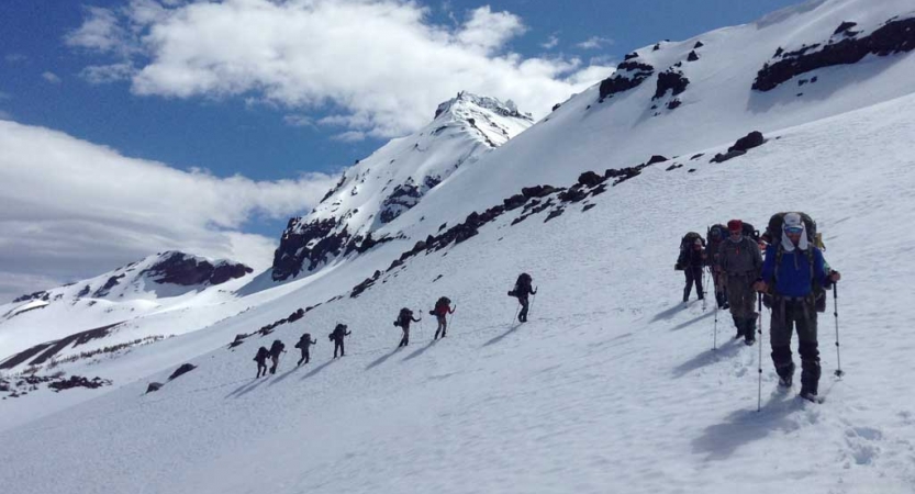 A line of people wearing snow gear, carrying backpacks and using trekking poles, hike through the snow of a mountainous landscape.